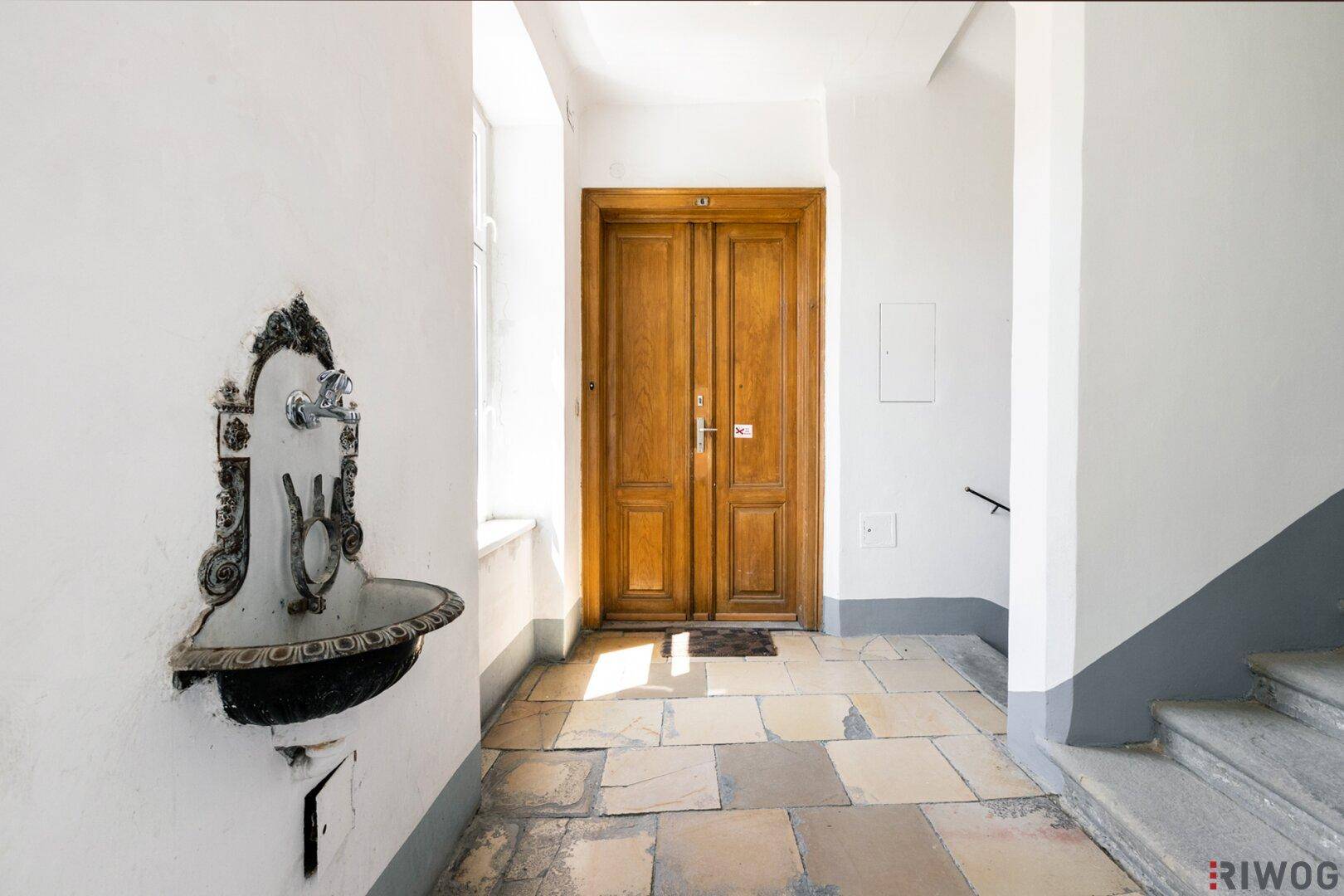 Heller Hausflur mit Steinfliesenboden, einer Holztür und einem dekorativen Wandbrunnen.