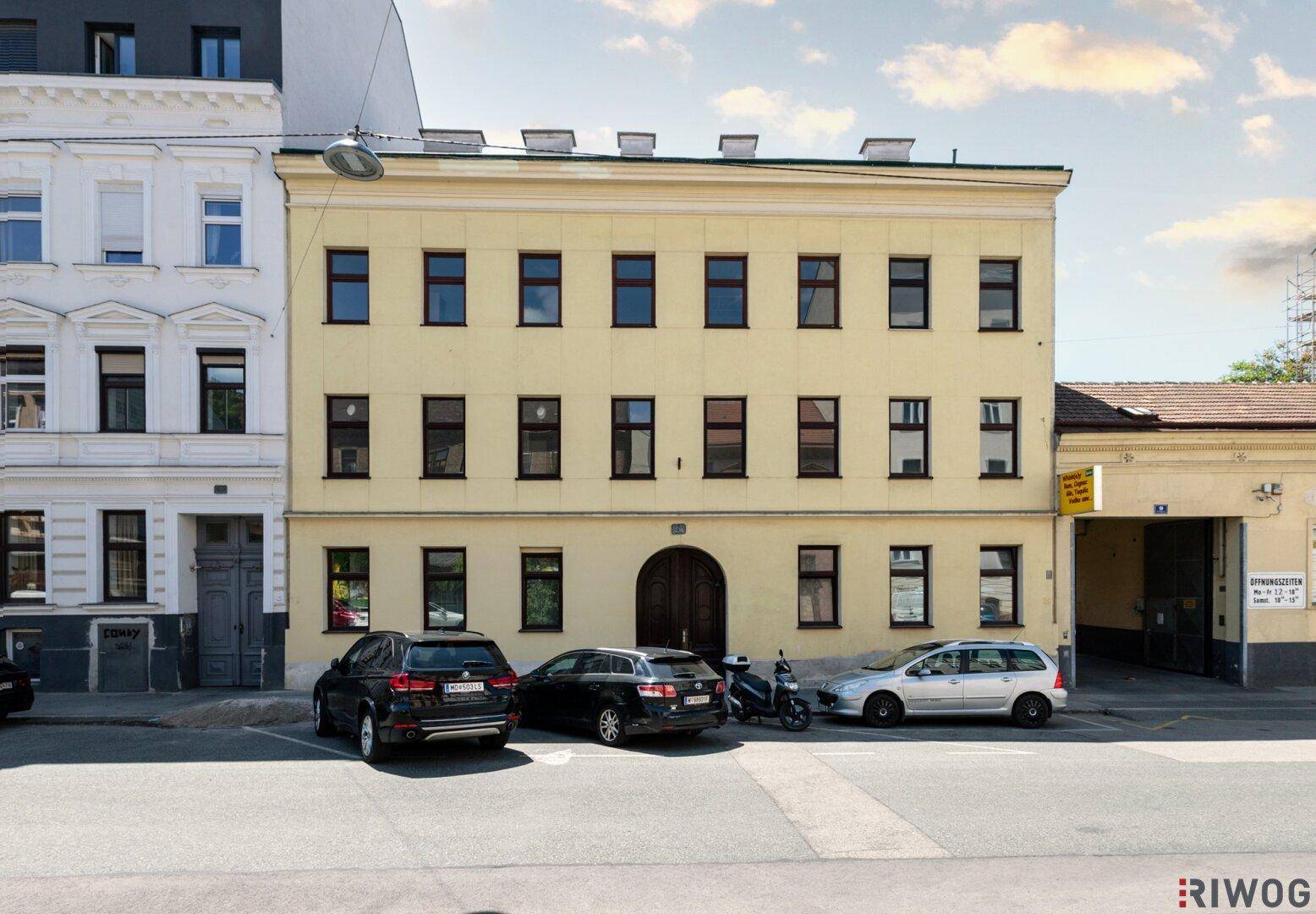 Gelbe Fassade eines Altbaus mit braunen Fenstern und parkenden Autos davor unter bewölktem Himmel.