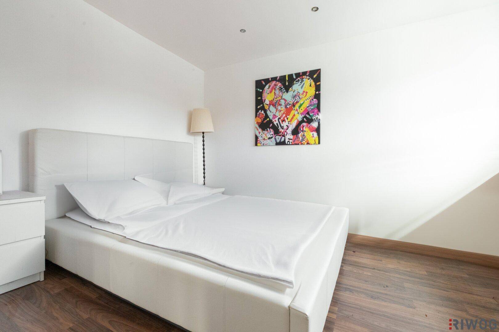 Helles Schlafzimmer mit weißem Bett, Holzboden und farbenfrohem Kunstwerk an der Wand.