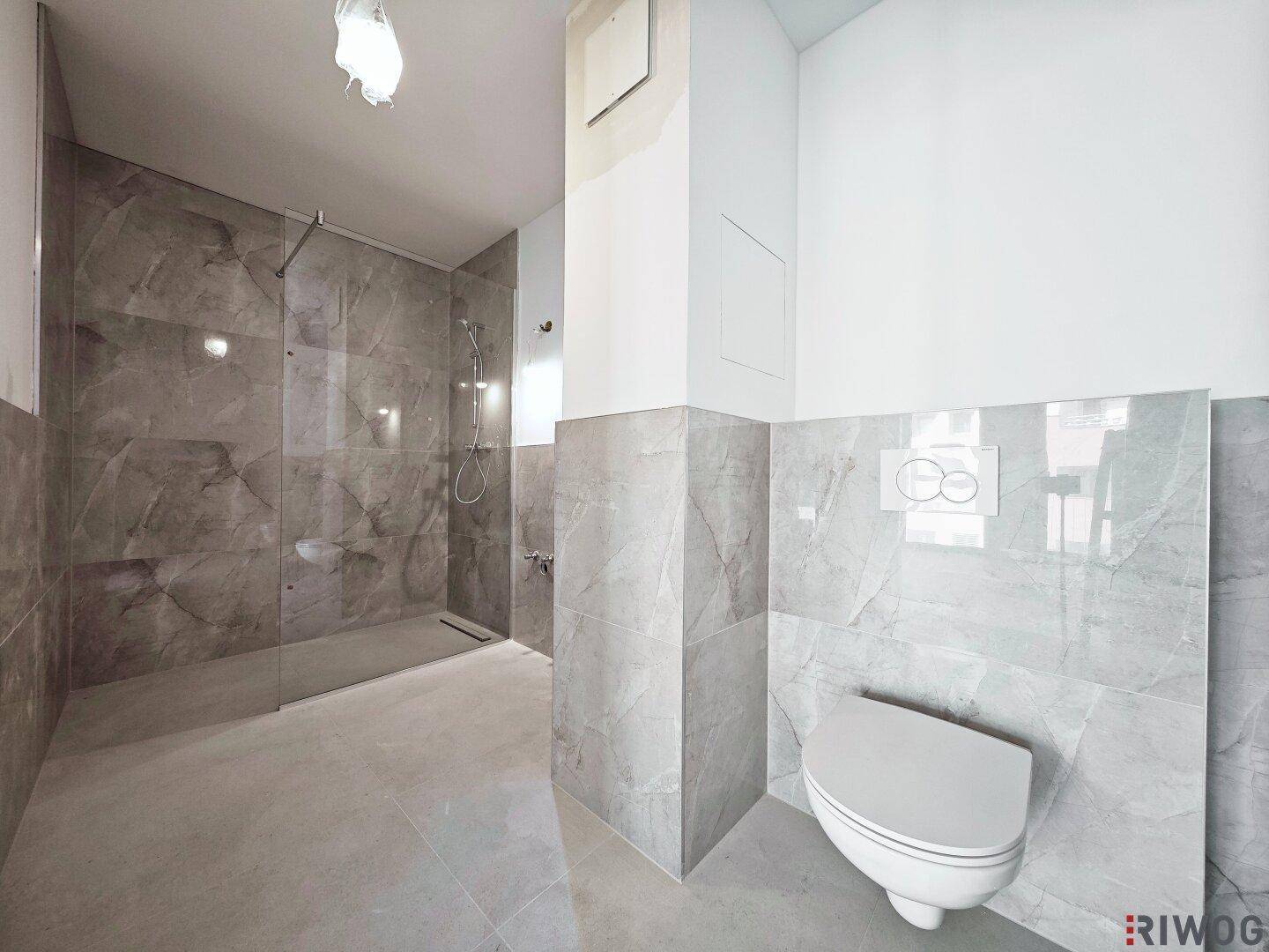 Modernes Badezimmer mit großformatigen grauen Fliesen, bodengleicher Dusche und Wand-WC.