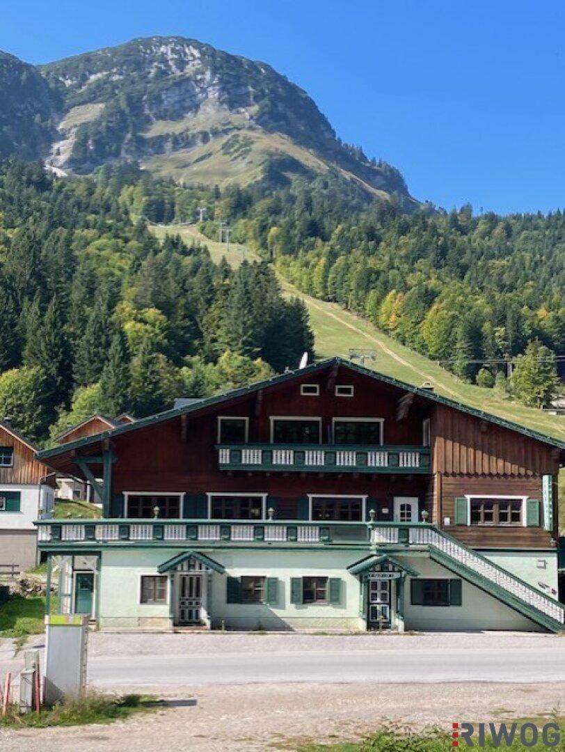 Alpines Gebäude mit Holzbalkonen und grünen Fensterläden vor einer beeindruckenden Bergkulisse.