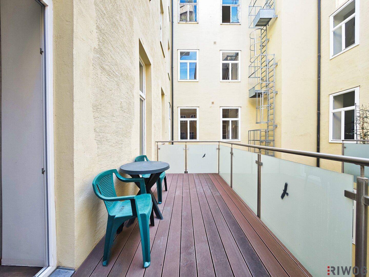 Geräumiger Balkon mit Holzboden und Sitzgelegenheiten, mit Blick auf den Innenhof und eine Feuertreppe.