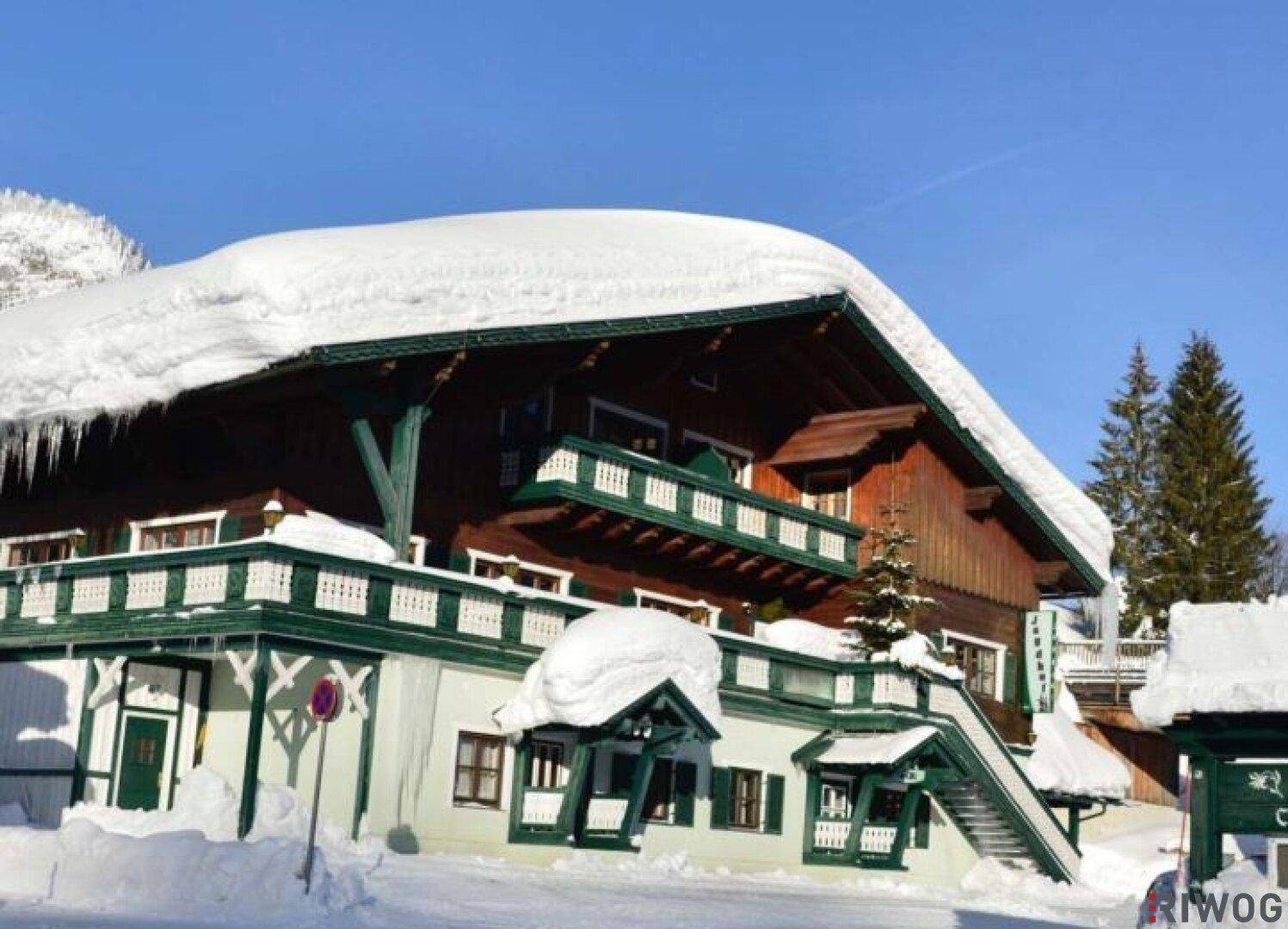 Winterliche Ansicht eines traditionellen Holzhauses mit schneebedecktem Dach und Eiszapfen.