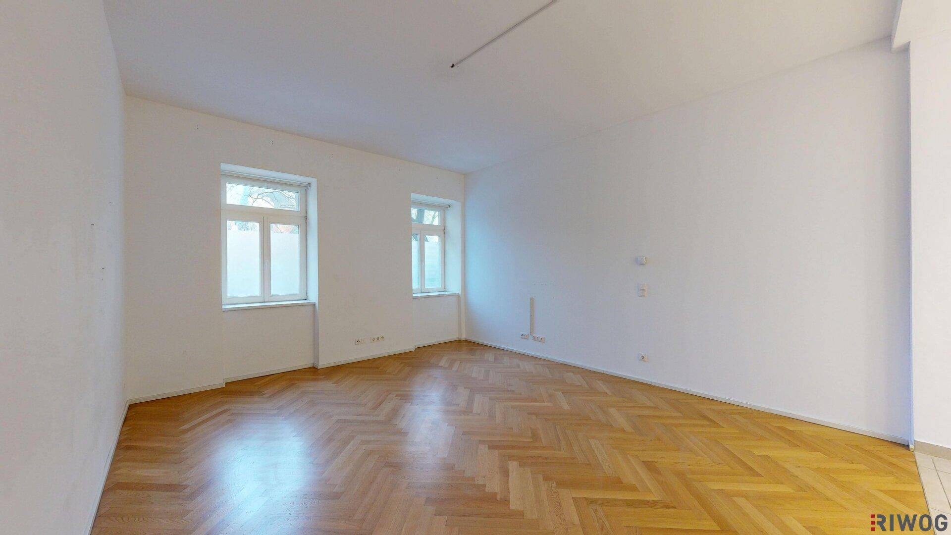 Geräumiger, leerer Raum mit Fischgrätparkett und zwei großen Fenstern, ideal als Wohnbereich.