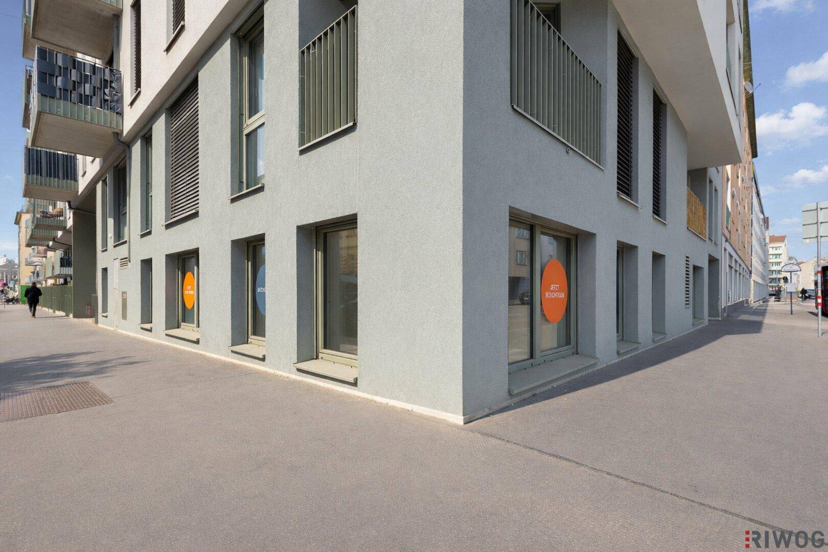 Moderne Gebäudeecke mit großen Schaufenstern im Erdgeschoss an einer belebten Straße bei Tageslicht.