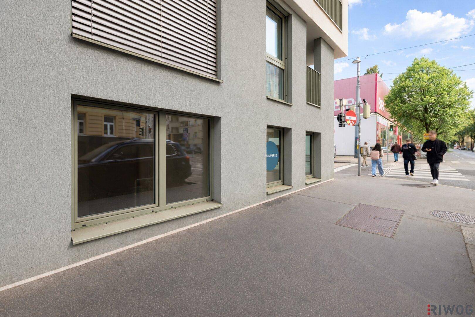 Fassade eines modernen Gebäudes mit großen Fenstern und Blick auf die belebte Straße und Bäume.