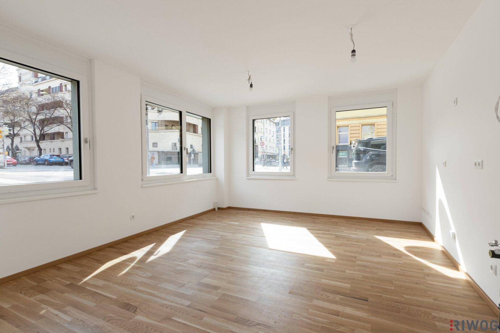 Heller, leerer Raum mit großen Fenstern, die viel Tageslicht hereinlassen und Holzboden.