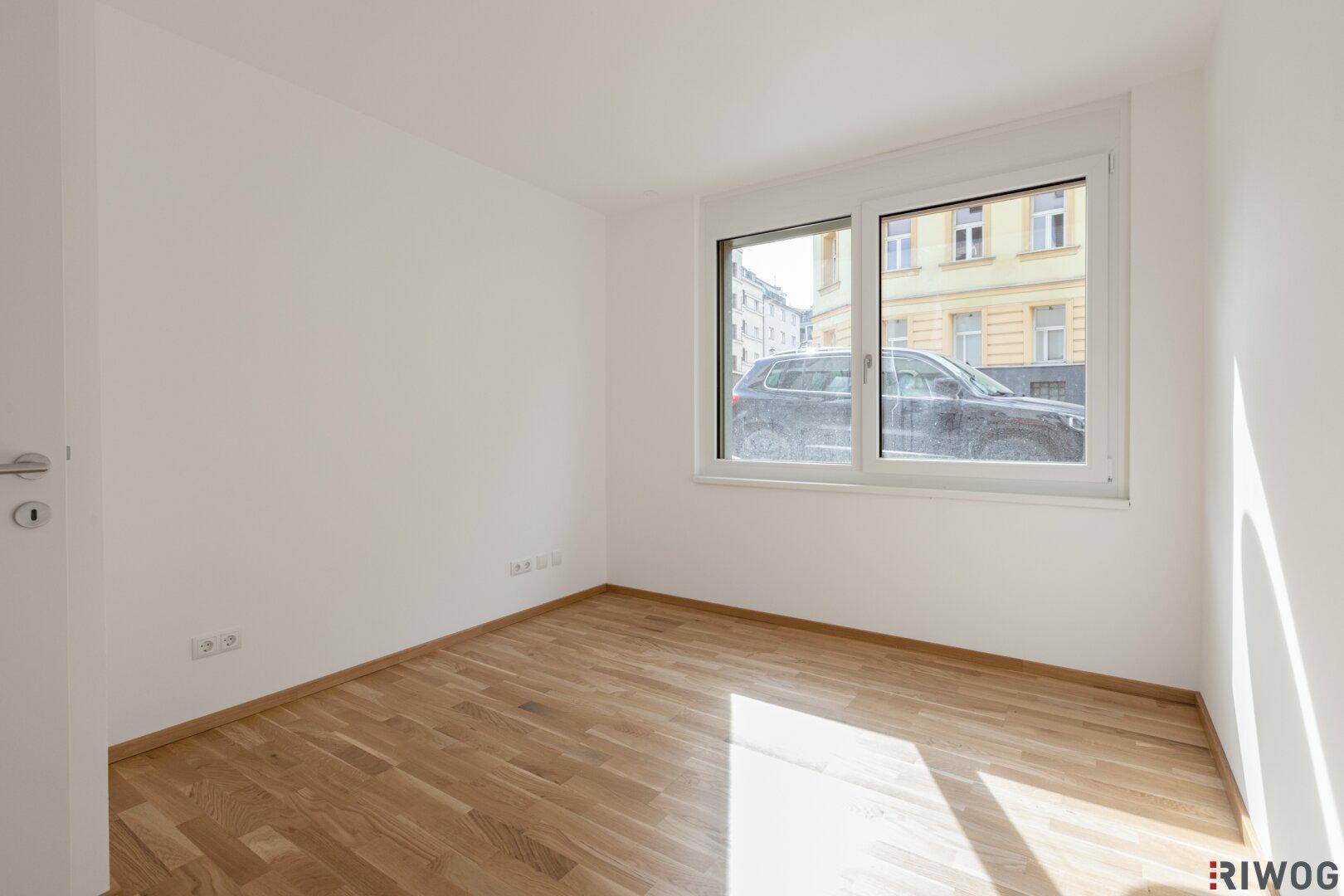 Heller, leerer Raum mit Holzboden und einem breiten Fenster, das viel Licht spendet.