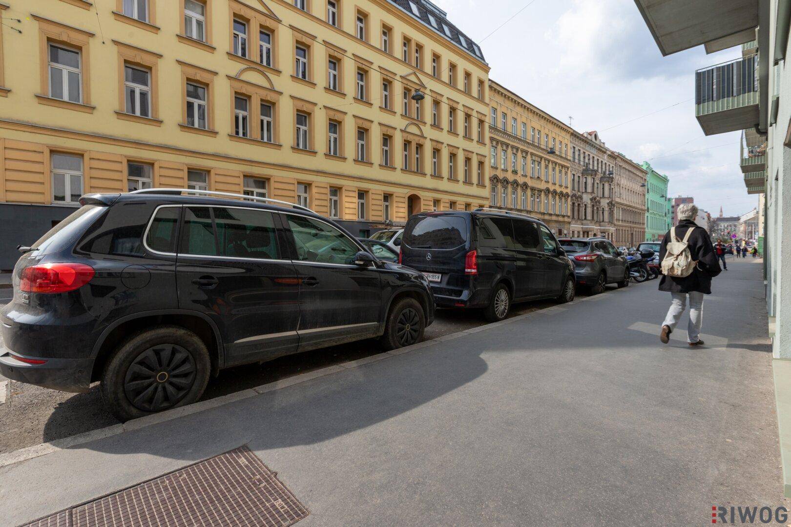 Blick auf eine belebte Straße mit parkenden Autos und einem historischen Gebäude im Hintergrund.