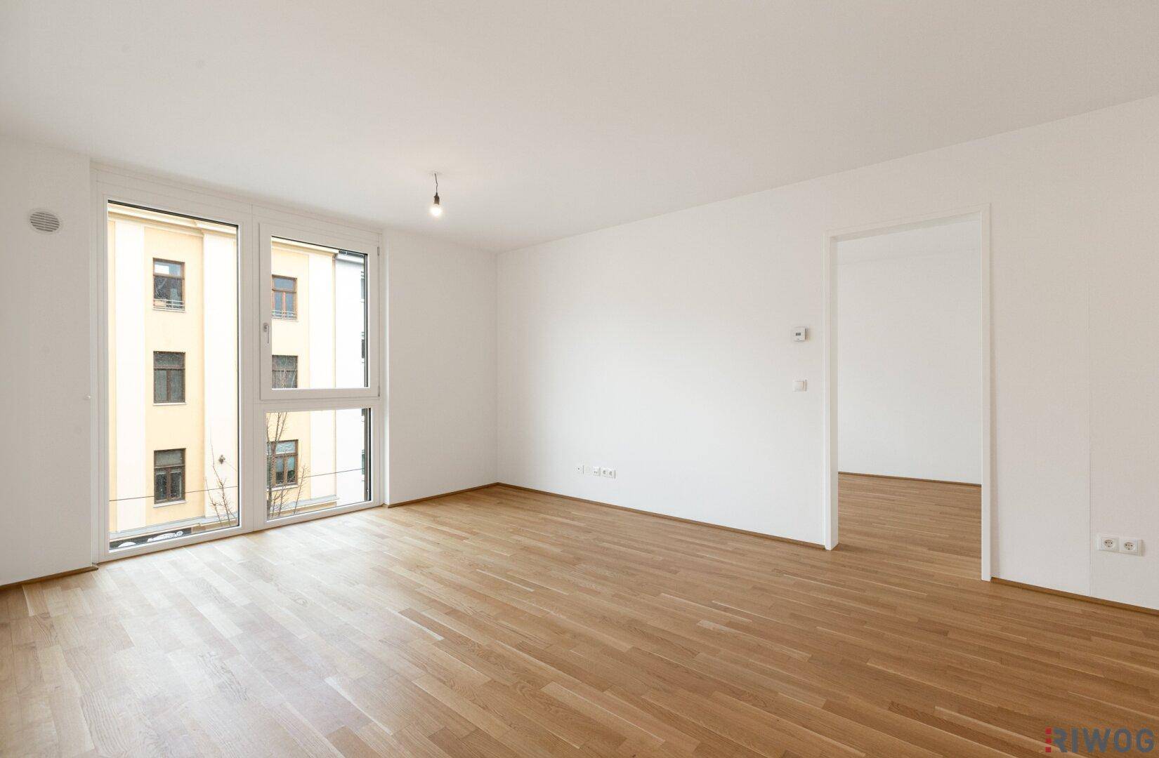 Geräumiges, leeres Wohnzimmer mit großen Fenstern und hellem Parkettboden.
