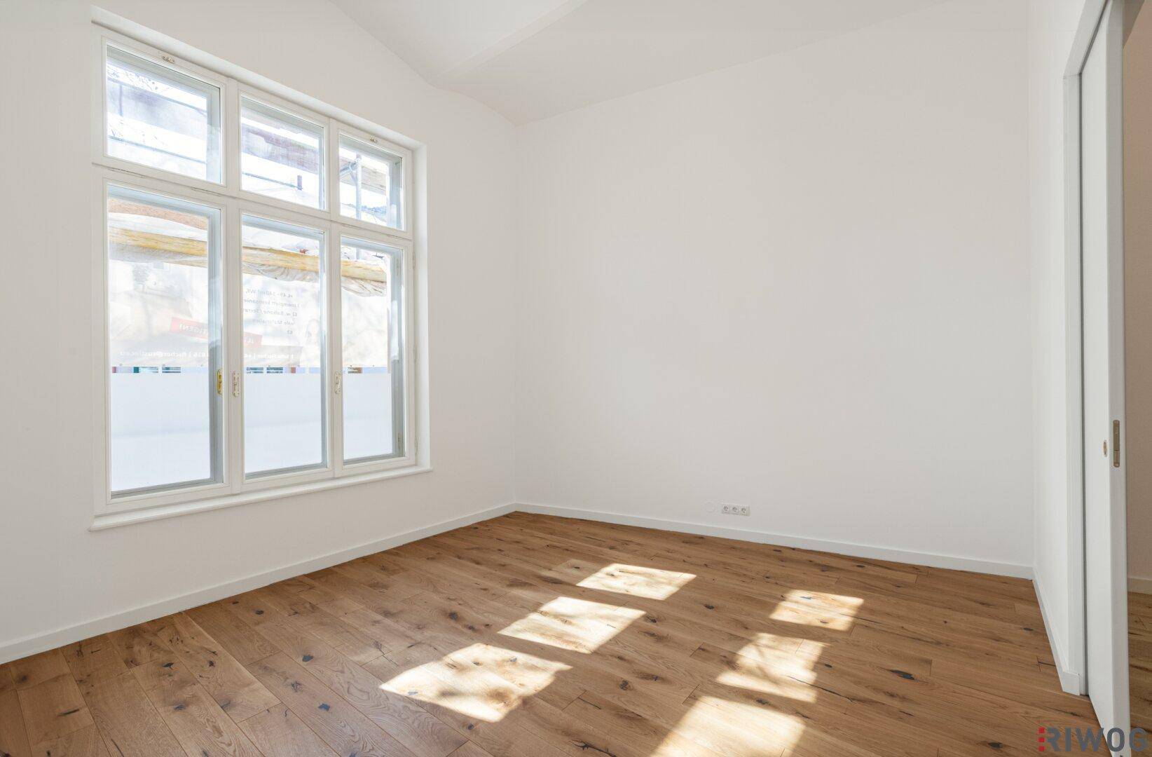 Heller Raum mit Holzboden und großen Fenstern, durch die Sonnenlicht auf den Boden fällt.