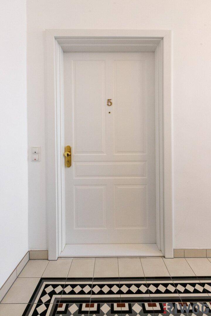 Weiße Wohnungstür mit goldener Klinke und dekorativem Fliesenboden im Eingangsbereich.