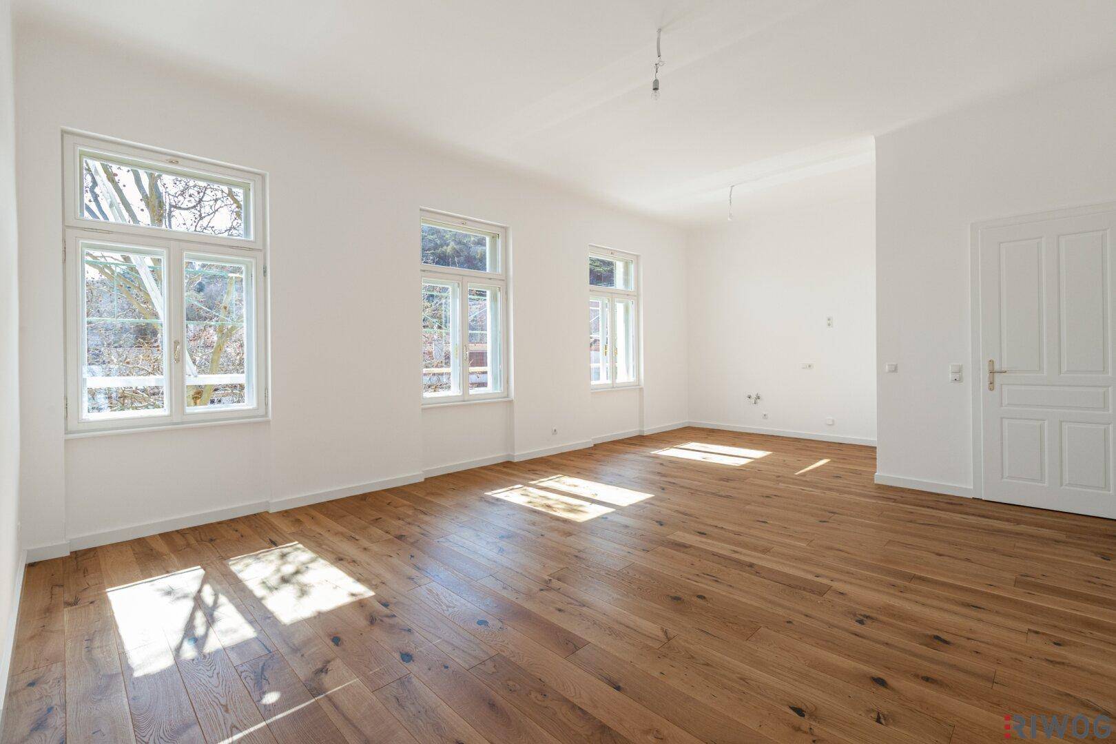 Lichtdurchfluteter, leerer Raum mit Holzboden und Blick ins Grüne durch die Fenster.