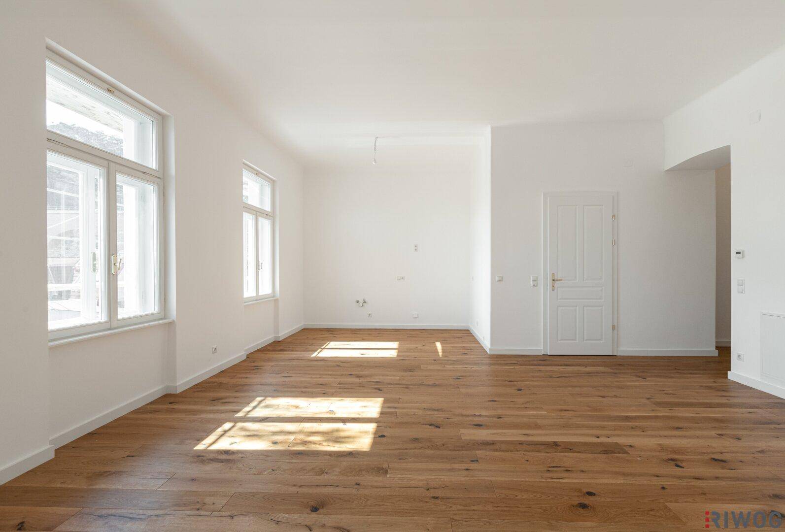 Großzügiger, leerer Raum mit Holzboden und hohen Fenstern, ideal für einen Wohnbereich.