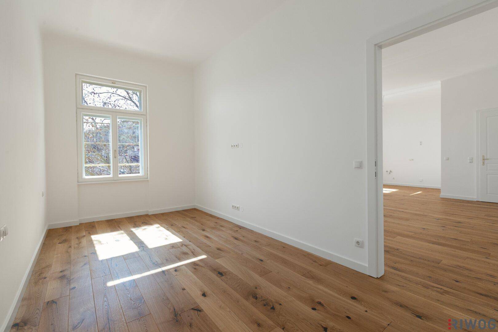 Helles, leeres Zimmer mit Holzboden und einem großen Fenster, das viel Tageslicht hereinlässt.