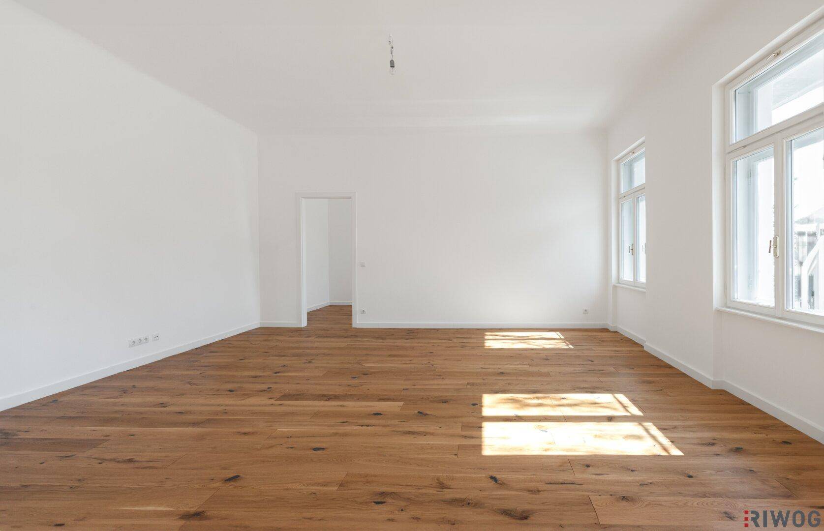 Geräumiges, leeres Wohnzimmer mit hellem Holzparkett und großen Fenstern, die viel Tageslicht hereinlassen.