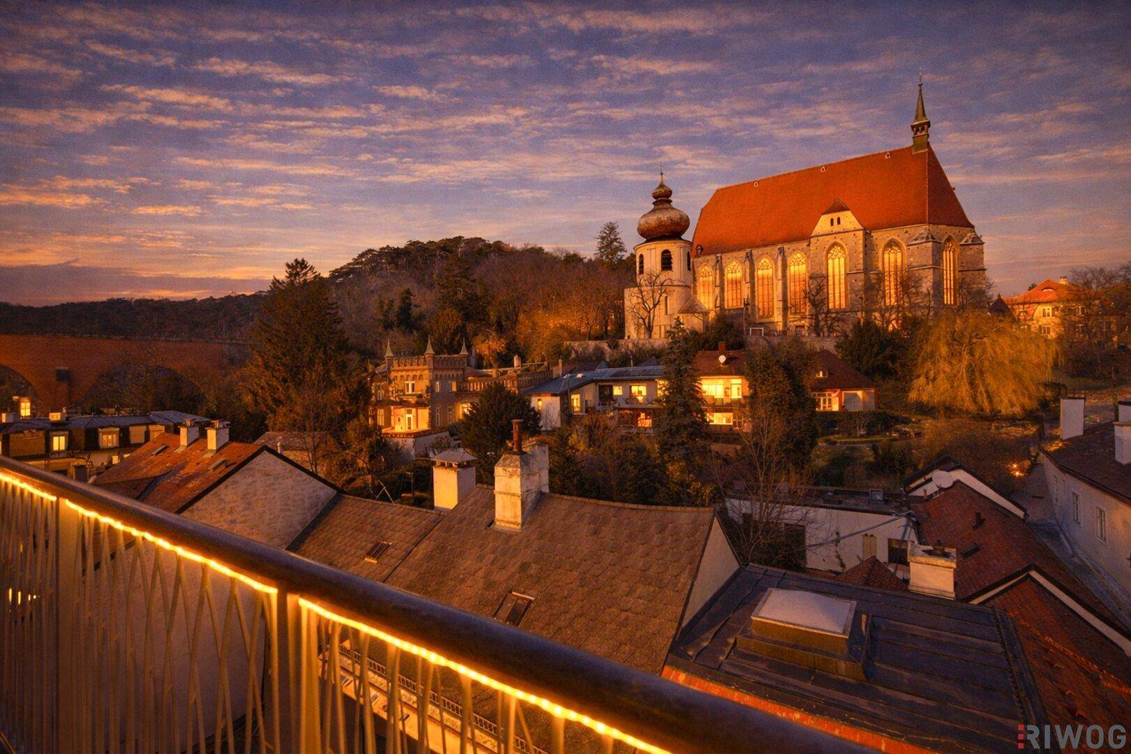 Malerischer Blick über die Dächer einer historischen Stadt mit beleuchteter Kirche bei Sonnenuntergang.
