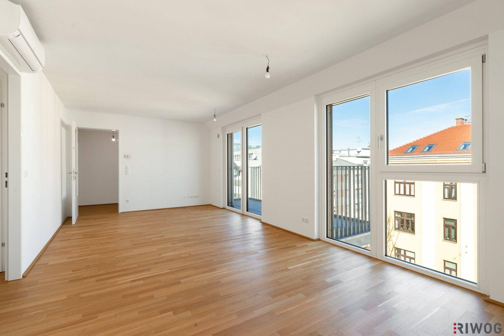 Helles, leeres Wohnzimmer mit Parkettboden, großen Fenstern und Zugang zum Balkon.