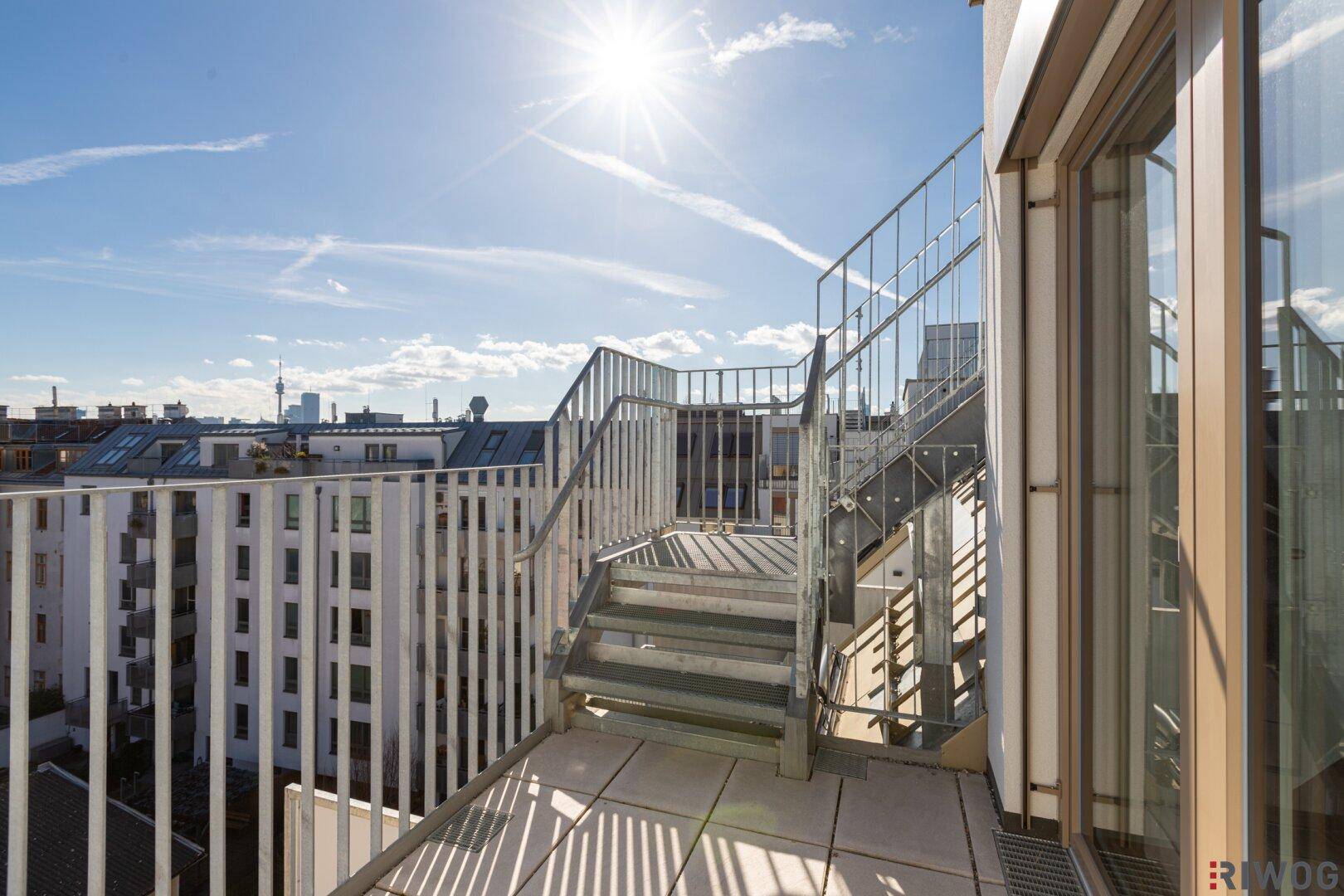 Sonniger Balkon mit Metallgeländer und Treppe, die zu einer oberen Terrasse führt.