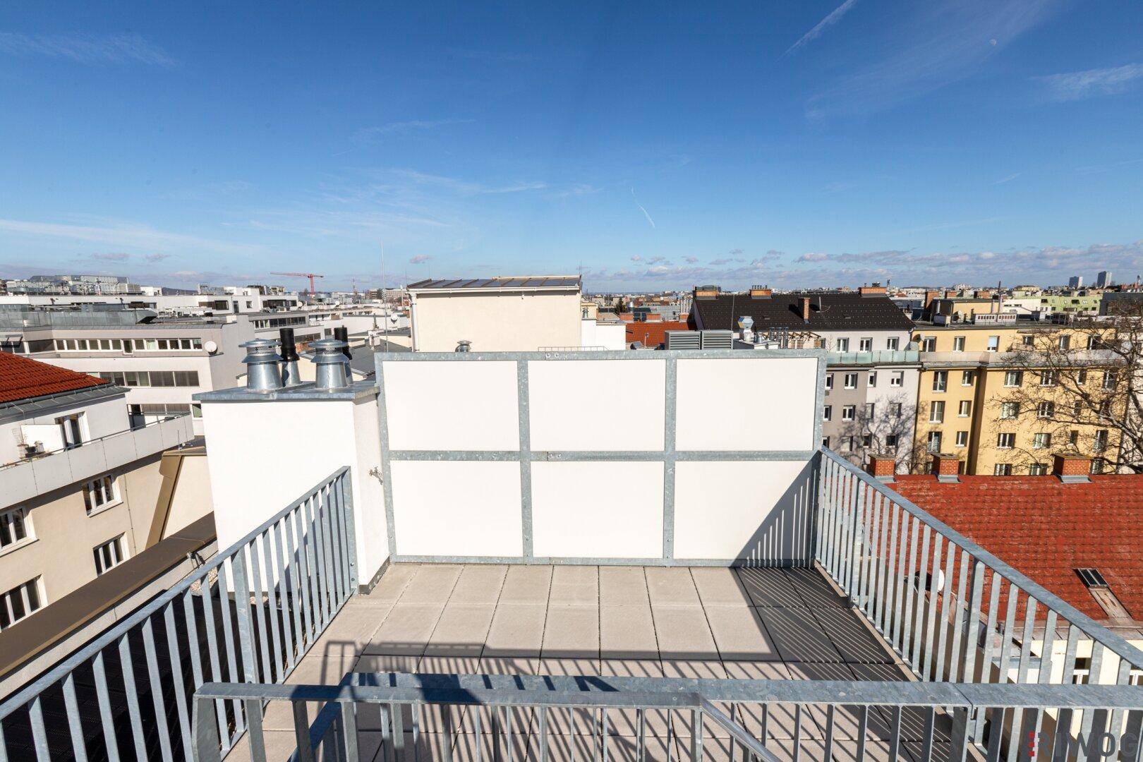 Unmöblierte Dachterrasse mit Blick auf die umliegenden Gebäude und den blauen Himmel.