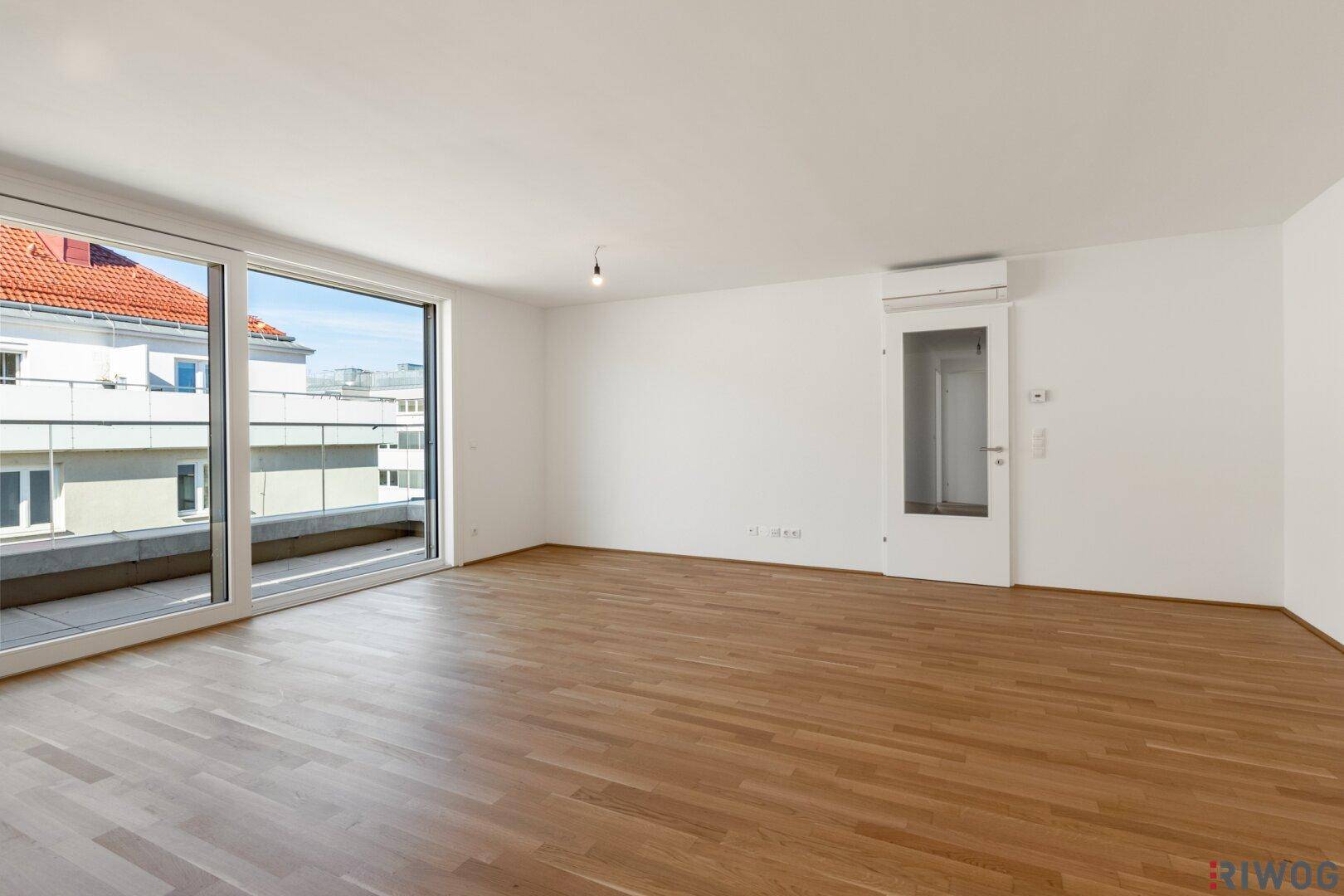 Helles, unmöbliertes Wohnzimmer mit Parkettboden und breiter Fensterfront zum Balkon.