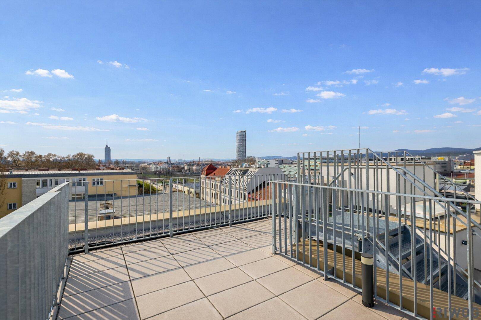 Geräumige Dachterrasse mit Fliesenboden und Panoramablick über die Stadt an einem sonnigen Tag.