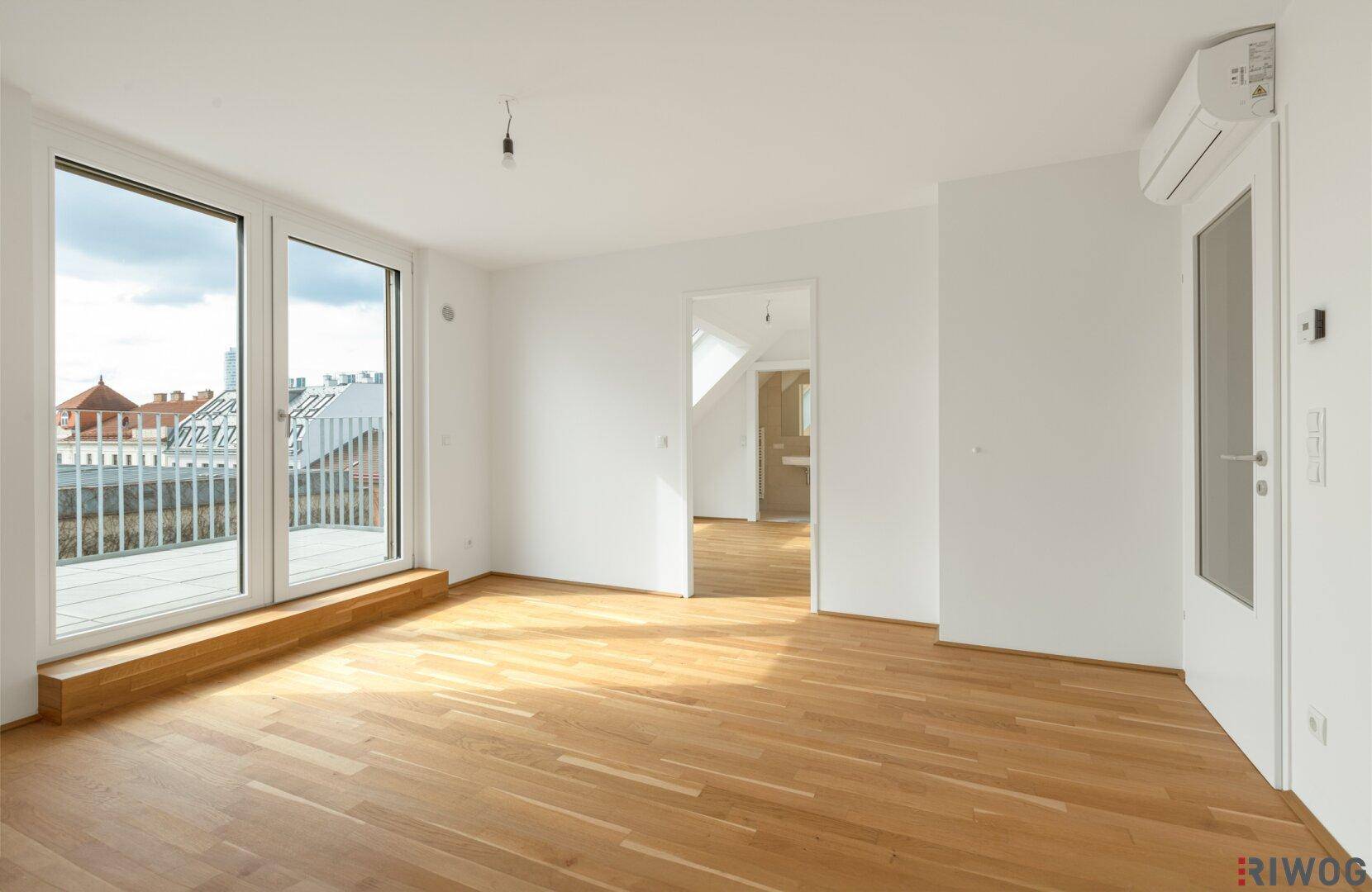 Heller, leerer Raum mit Holzboden und großen Fenstern, die Zugang zu einem Balkon bieten.