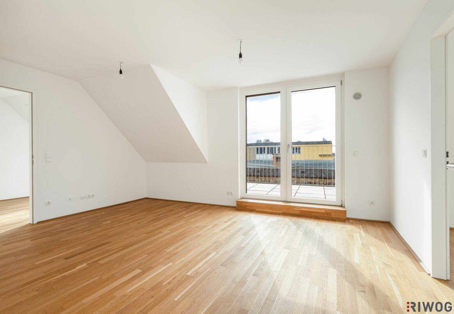 Geräumiger, heller Raum mit Holzboden, Schrägdecke und großen Fenstern zum Balkon.