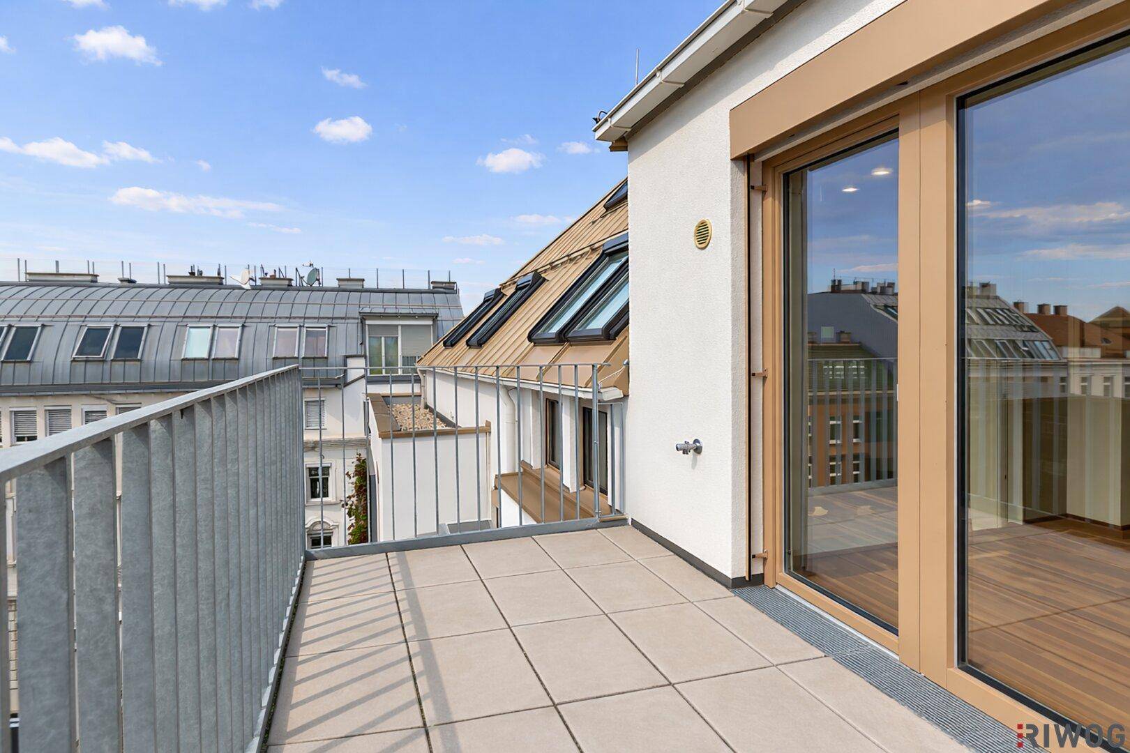 Heller Balkon mit Fliesenboden und Zugang zu einem Innenraum, umgeben von Dachlandschaften.