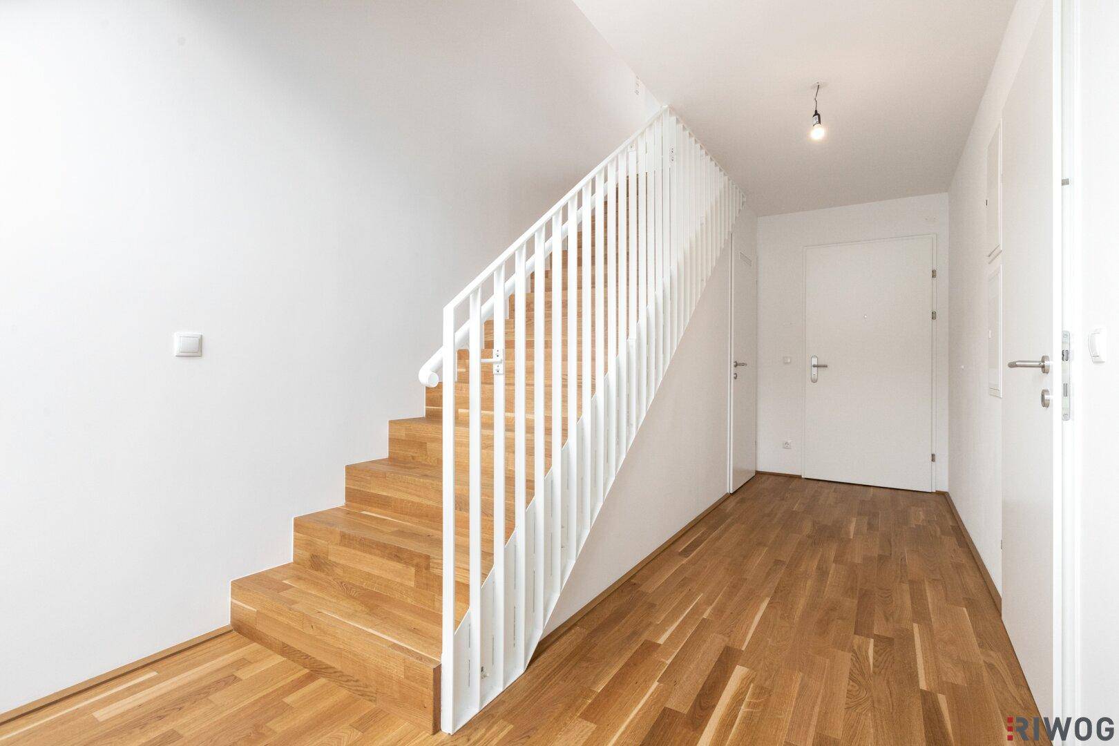 Heller Flur mit Holztreppe, weißem Geländer und Parkettboden.