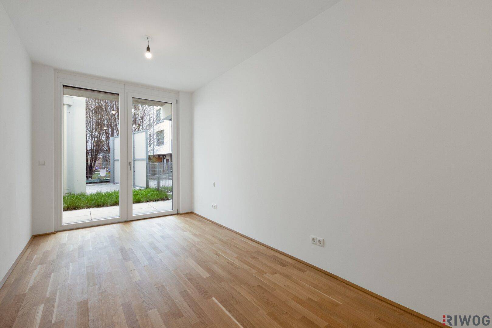 Leeres Zimmer mit hellem Parkettboden und großen Fenstern, die viel Tageslicht hereinlassen.