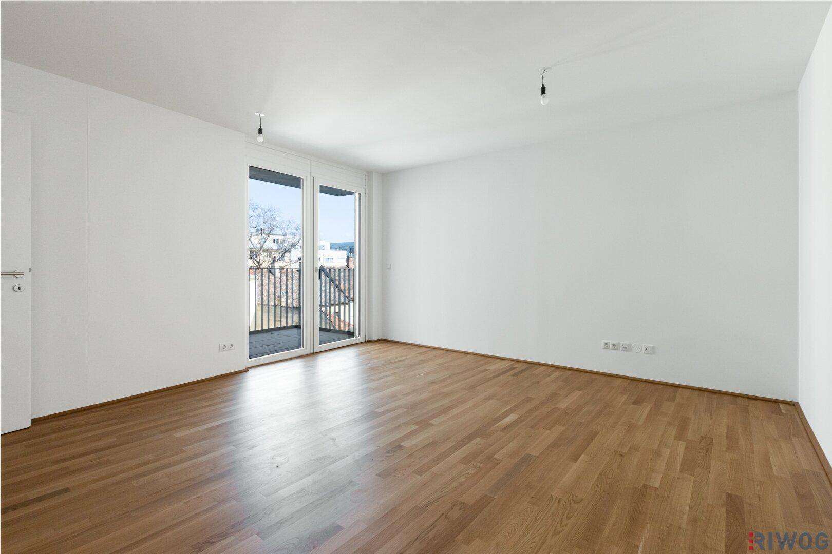 Helles Zimmer mit Holzboden und großen Fenstern, die viel Tageslicht hereinlassen.