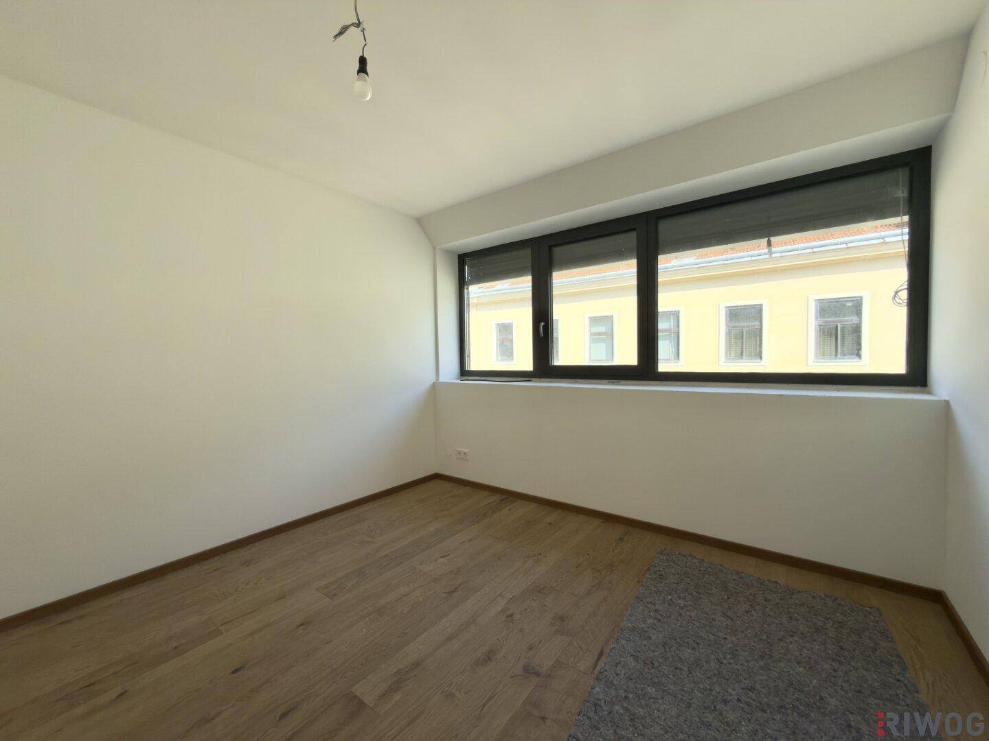 Weiteres helles Zimmer mit Holzboden und mehreren Fenstern, die viel Tageslicht hereinlassen.
