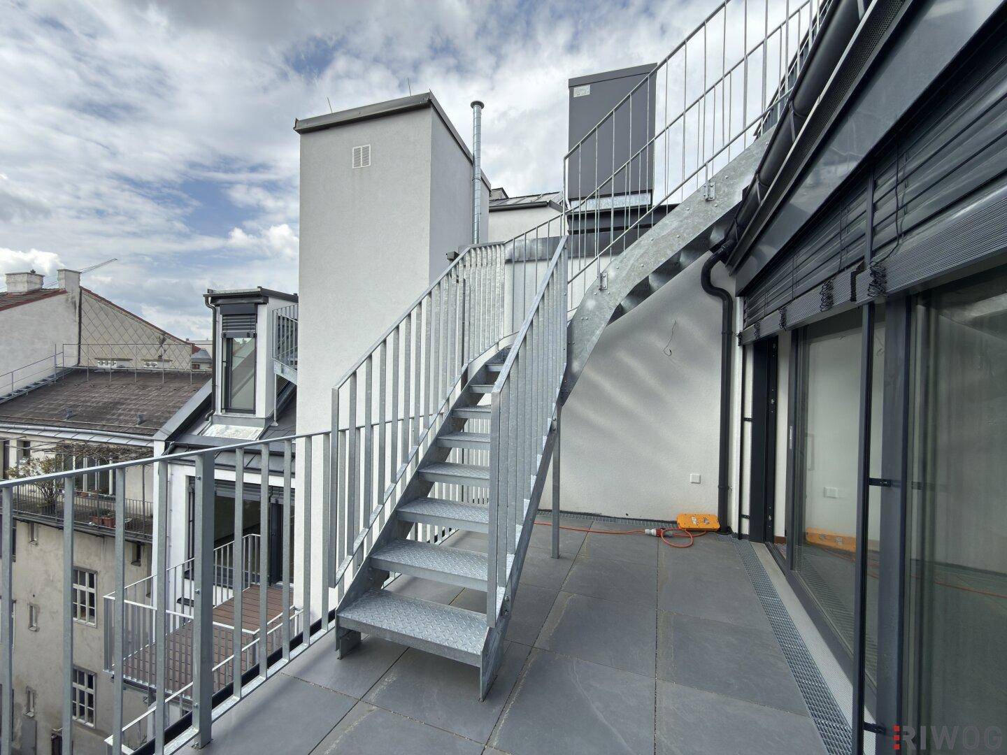 Außentreppe aus Metall auf einer Terrasse, die zu einer weiteren Ebene führt, mit Stadtblick.