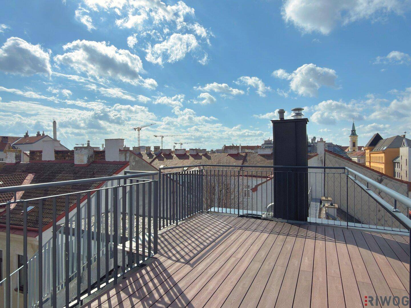 Sonnige Dachterrasse mit Holzboden und weitem Blick über die städtische Umgebung unter blauem Himmel.