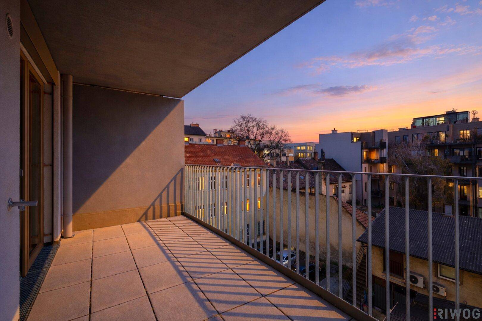 Geräumiger Balkon mit Fliesenboden und Blick auf die umliegenden Gebäude bei Sonnenuntergang.