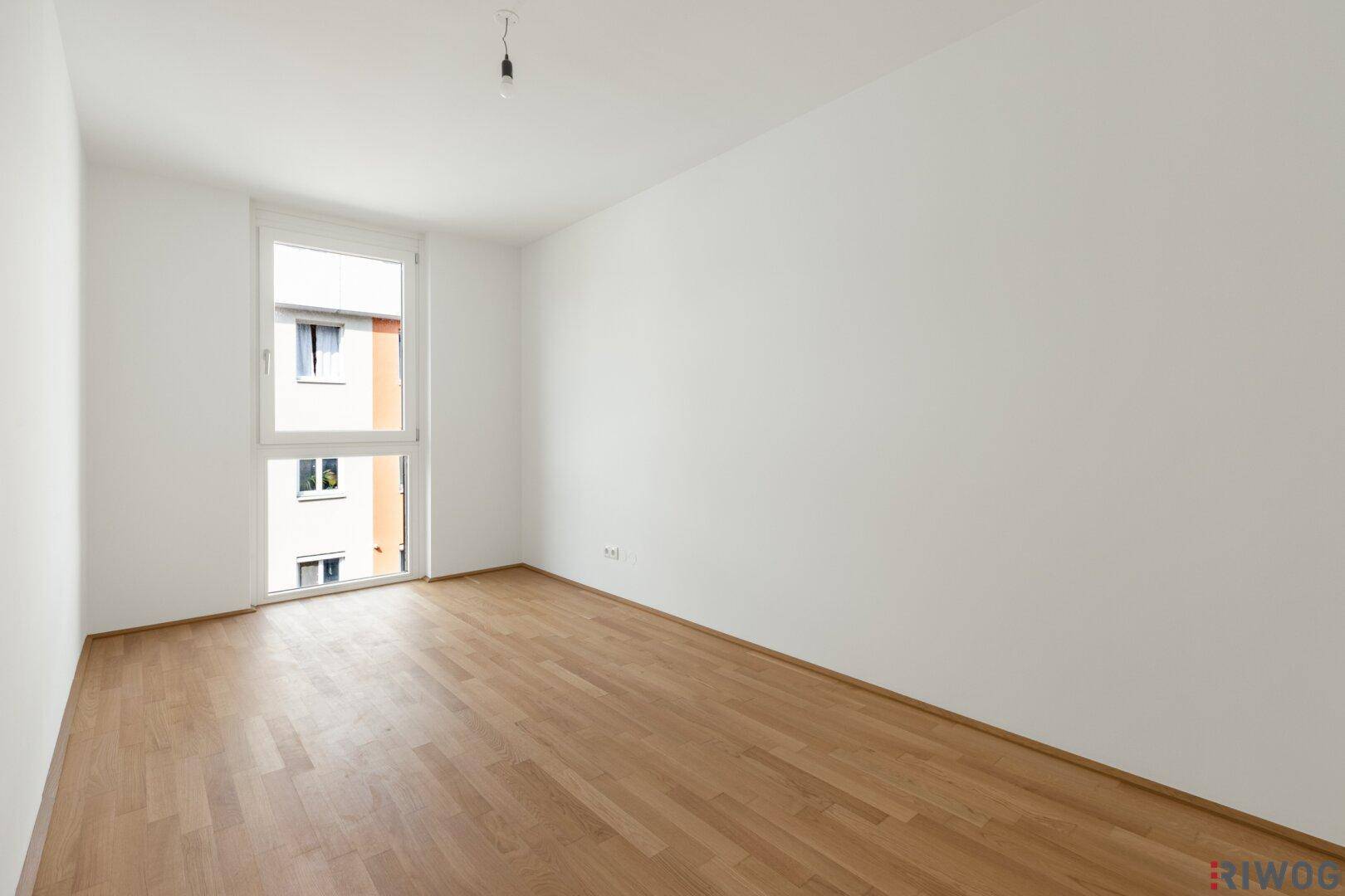 Helles, leeres Zimmer mit Parkettboden und großem Fenster, bietet viel Tageslicht.