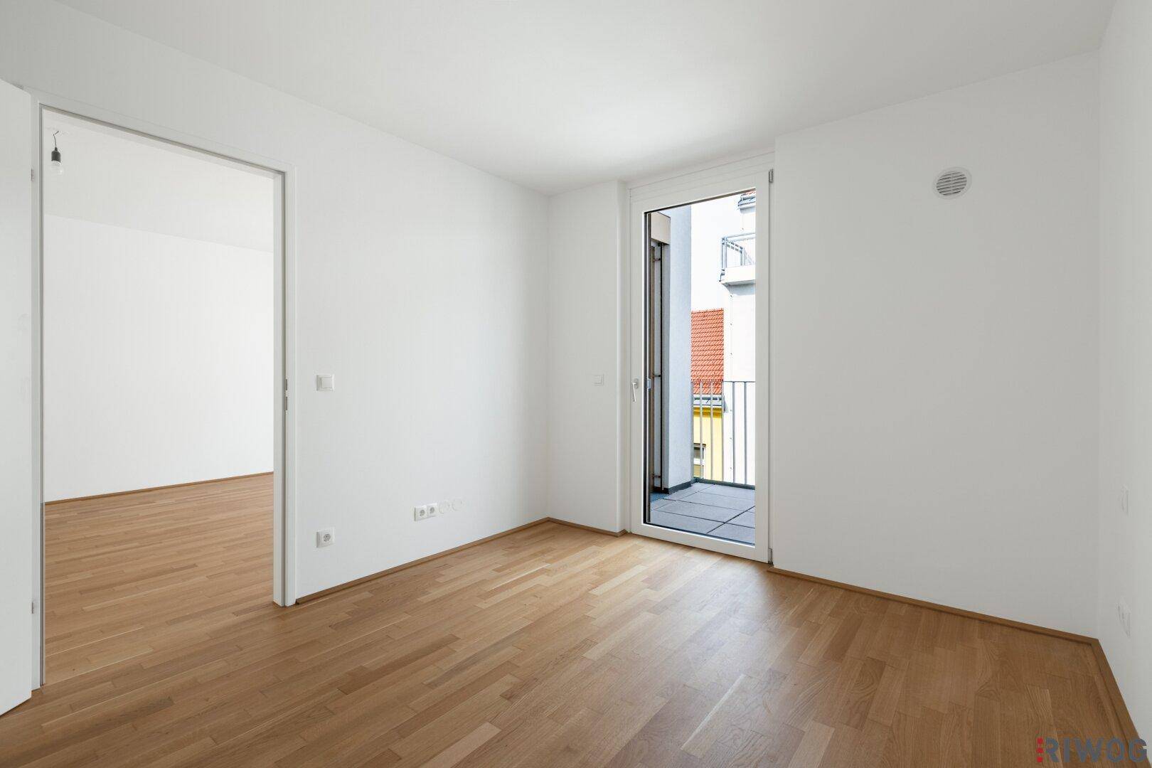 Helles Zimmer mit Parkettboden und Zugang zu einem kleinen Balkon.