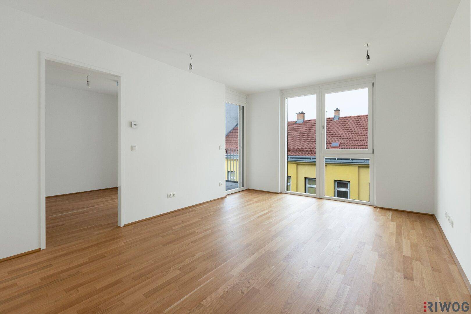 Helles, leeres Wohnzimmer mit Parkettboden und Zugang zum Balkon.