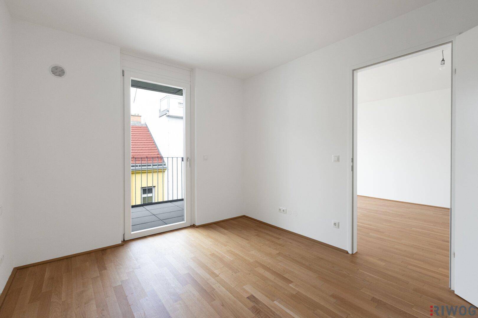 Leerer Raum mit Parkettboden und großer Balkontür, die viel Licht hereinlässt.