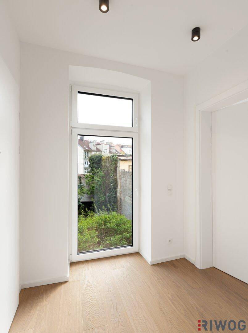 Heller Raum mit Holzboden und einem hohen Fenster, das einen grünen Ausblick auf Pflanzen bietet.