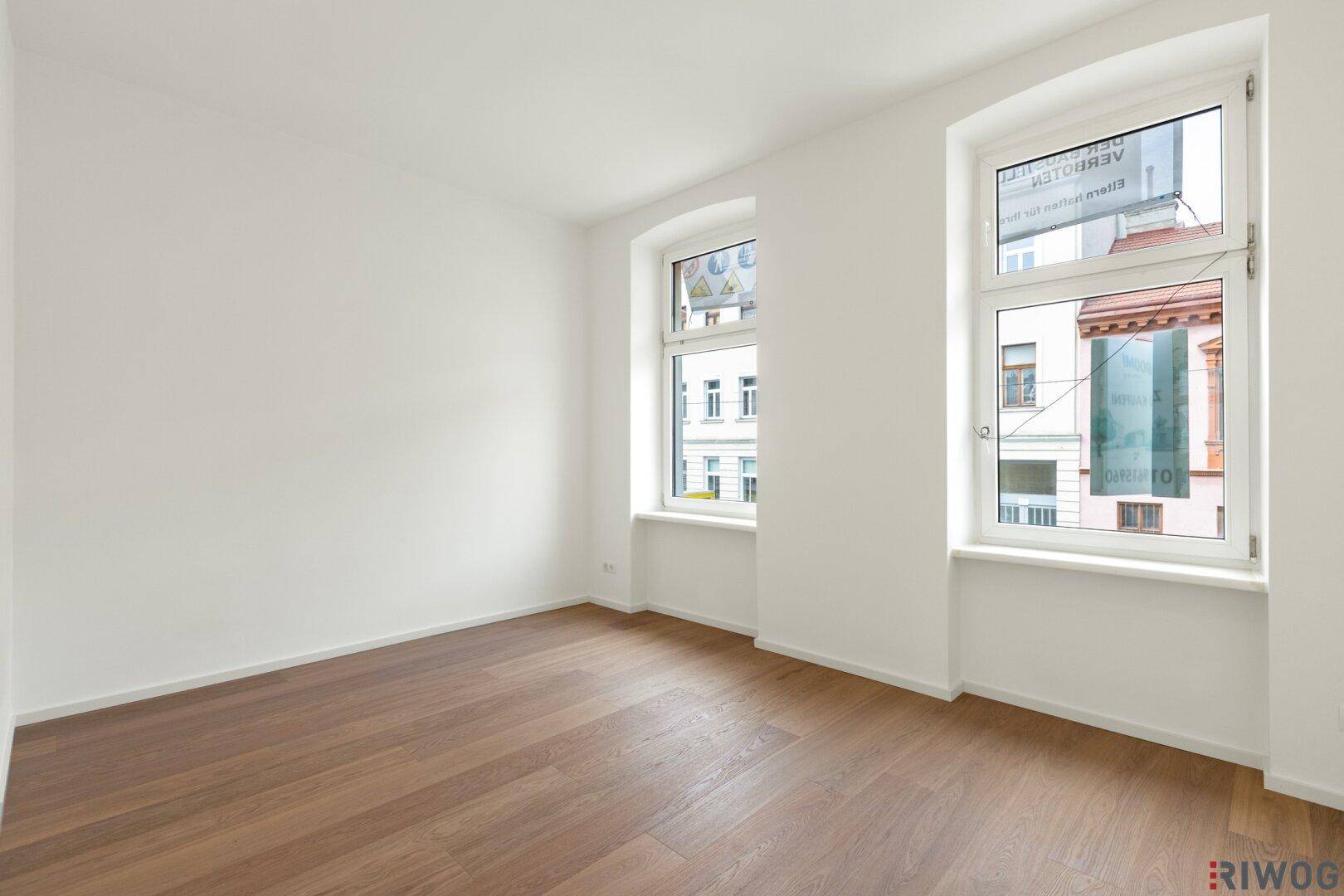 Heller Raum mit Holzboden und zwei Fenstern, die einen Blick auf die umliegenden Gebäude bieten.