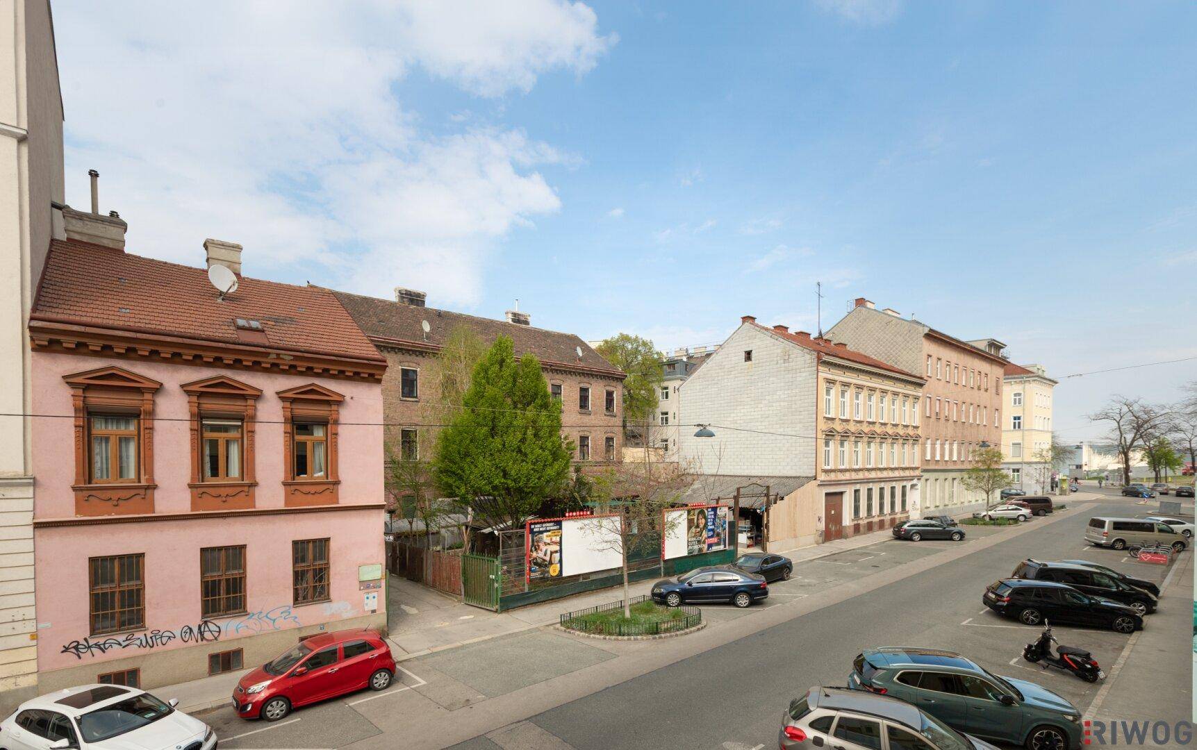 Blick auf eine belebte Stadtstraße mit parkenden Autos und historischen Gebäuden unter blauem Himmel.
