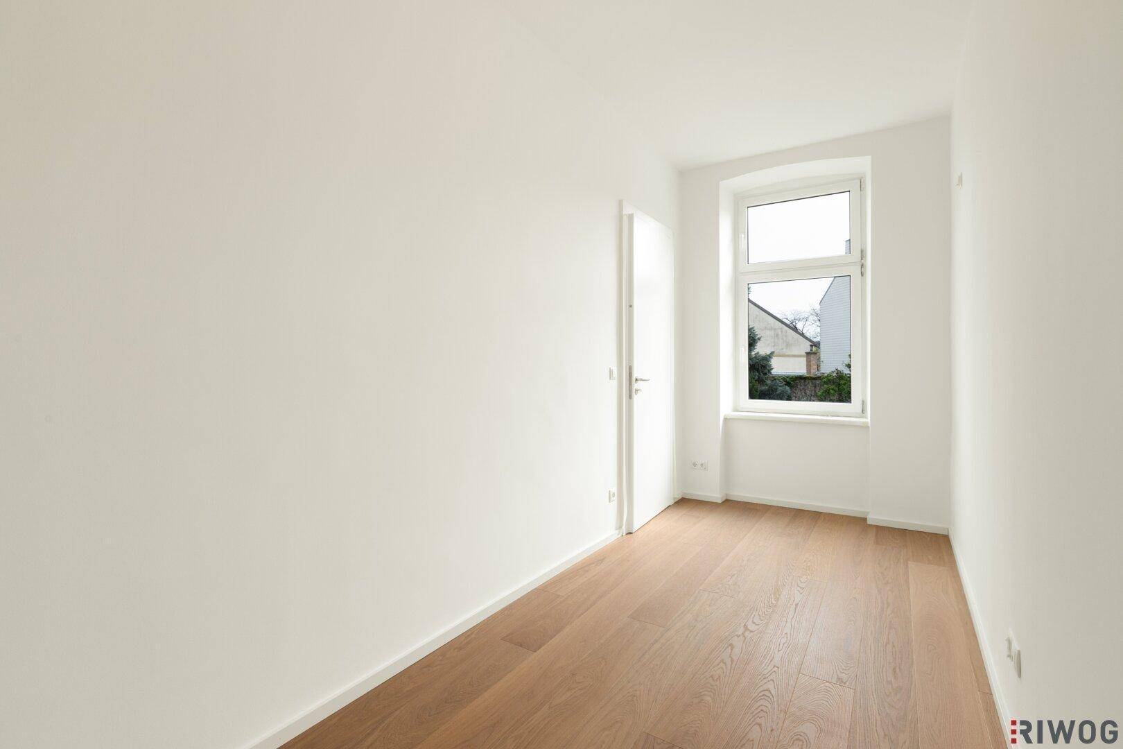 Leerer Raum mit hellem Holzboden, weißen Wänden und einem Fenster, das Tageslicht hereinlässt.