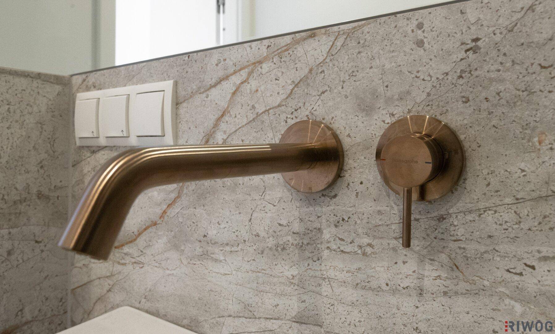 Detailaufnahme eines modernen, wandmontierten Wasserhahns in Bronzeoptik vor einer grauen Marmorwand.