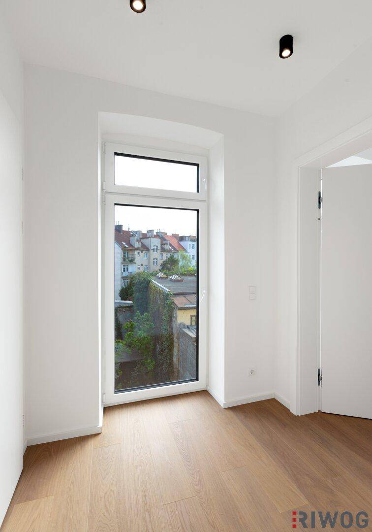 Helles Zimmer mit Holzboden und einem hohen Fenster, das einen Blick auf die umliegenden Gebäude bietet.