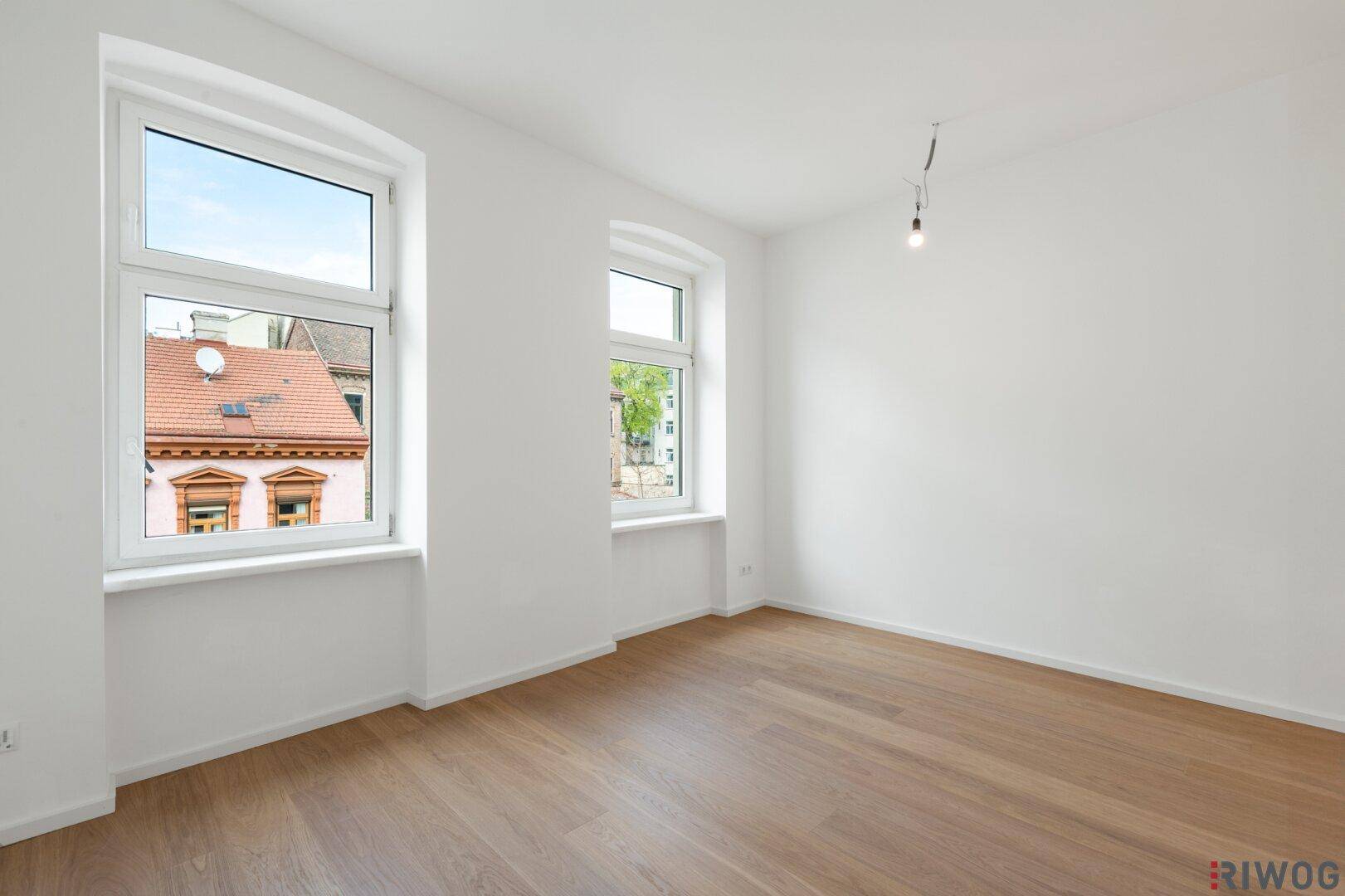 Helles, leeres Zimmer mit Holzboden und zwei großen Fenstern, die einen Blick auf die Nachbarschaft bieten.