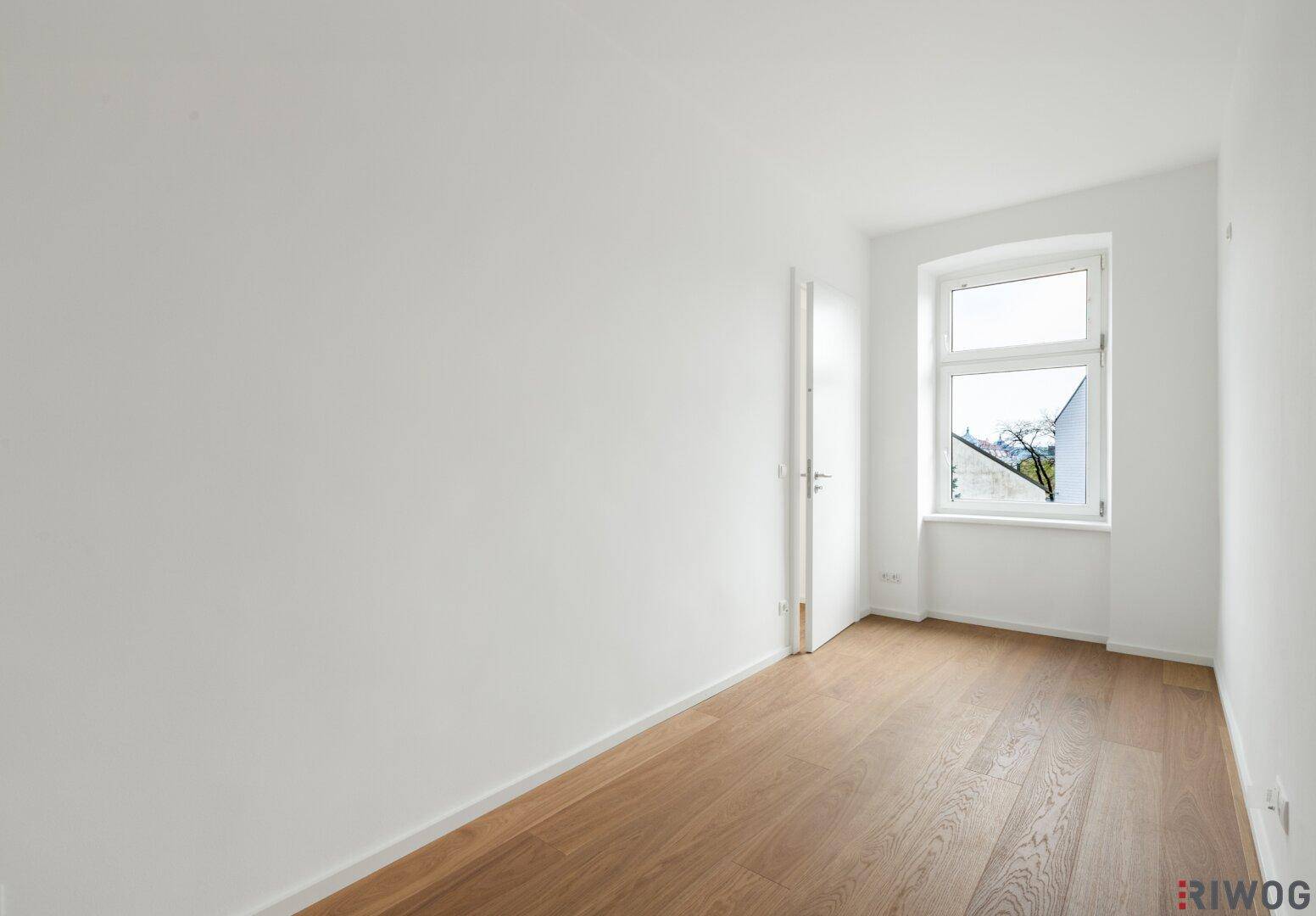 Leeres Schlafzimmer mit Holzboden, weiß gestrichenen Wänden und einem Fenster mit Blick ins Grüne.