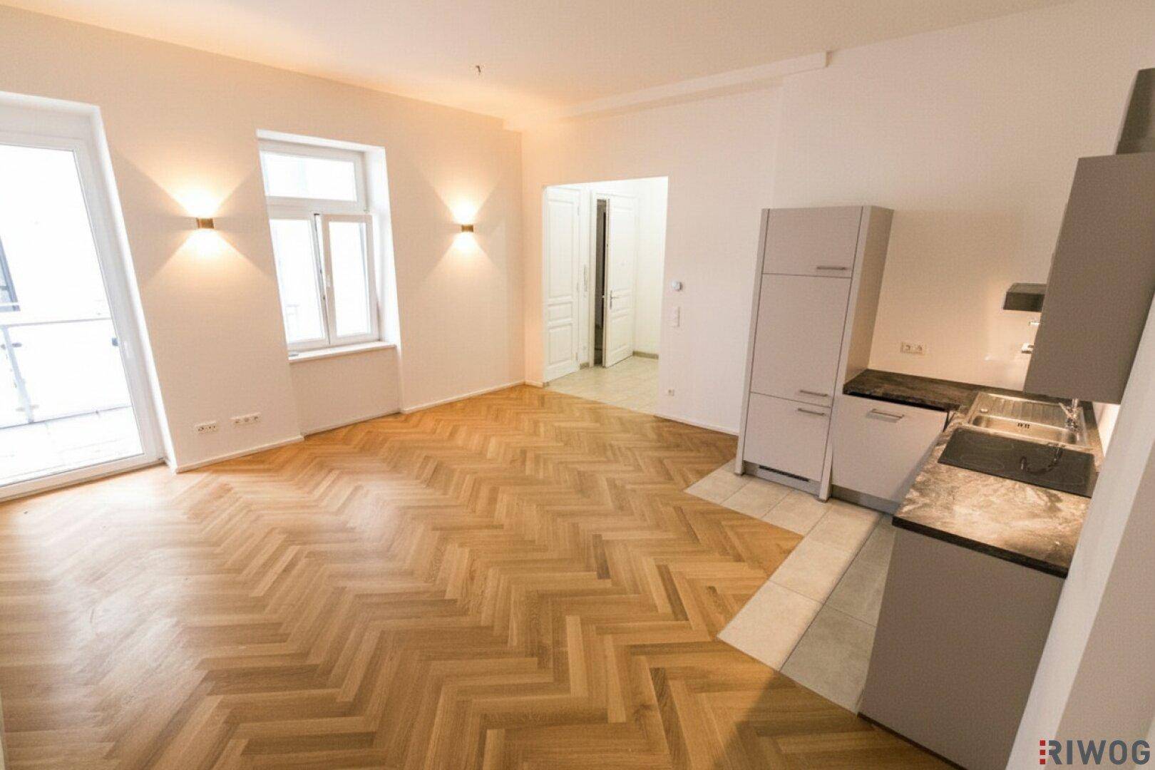 Leerer Raum mit Fischgrätparkett, großen Fenstern und vorbereiteten Küchenanschlüssen.