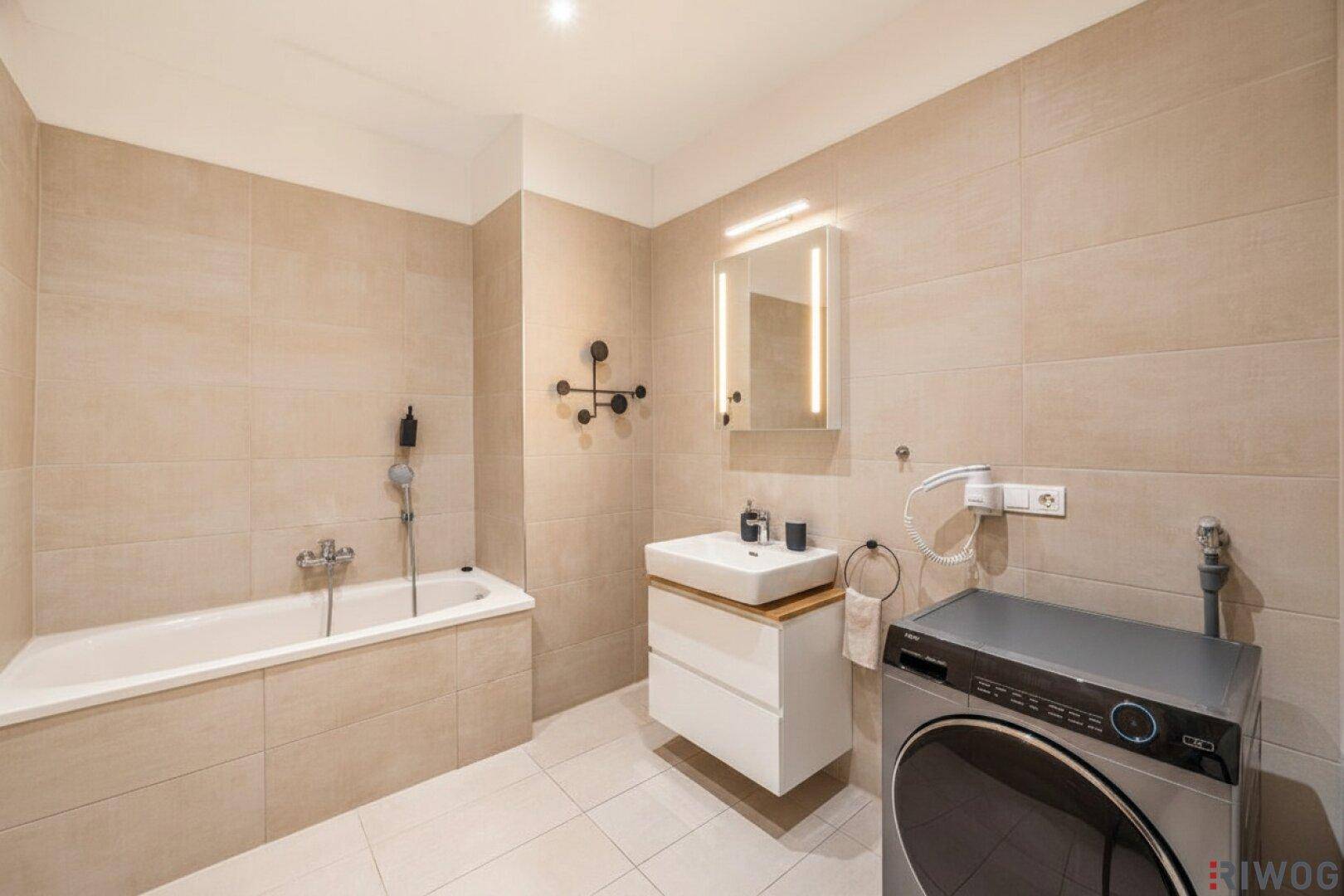 Modernes Badezimmer mit Badewanne, Waschbecken und integrierter Waschmaschine in hellen Fliesen.